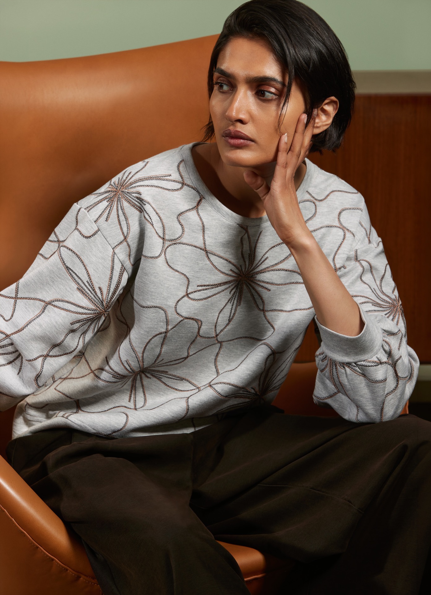Vrouw zit op leren stoel, draagt grijze trui van Atmos met bruine en beige bloemenprint, hand tegen oor, nadenkende blik.